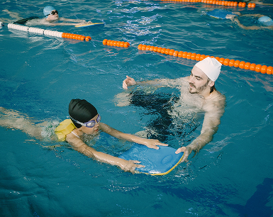 Schwimmunterricht: Erwachsener begleitet ein Kind mit Schwimmbrett im Becken